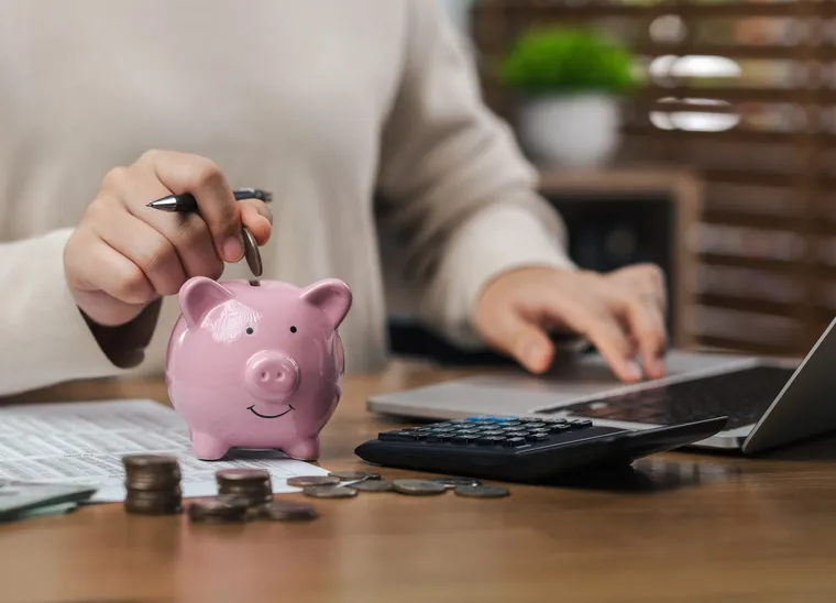 A person dropping a coin into a piggy bank while working at a laptop beside a calculator and financial paperwork