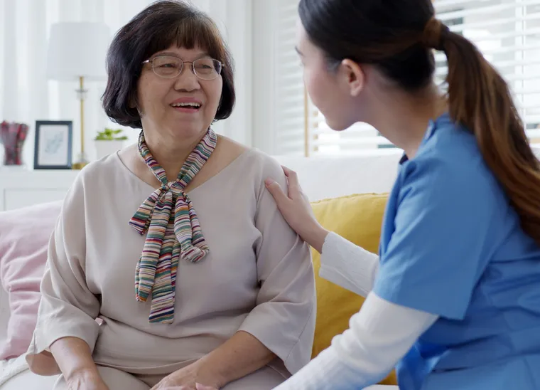 A caregiver in blue scrubs sitting beside a smiling older woman on a sofa, a hand resting on her shoulder